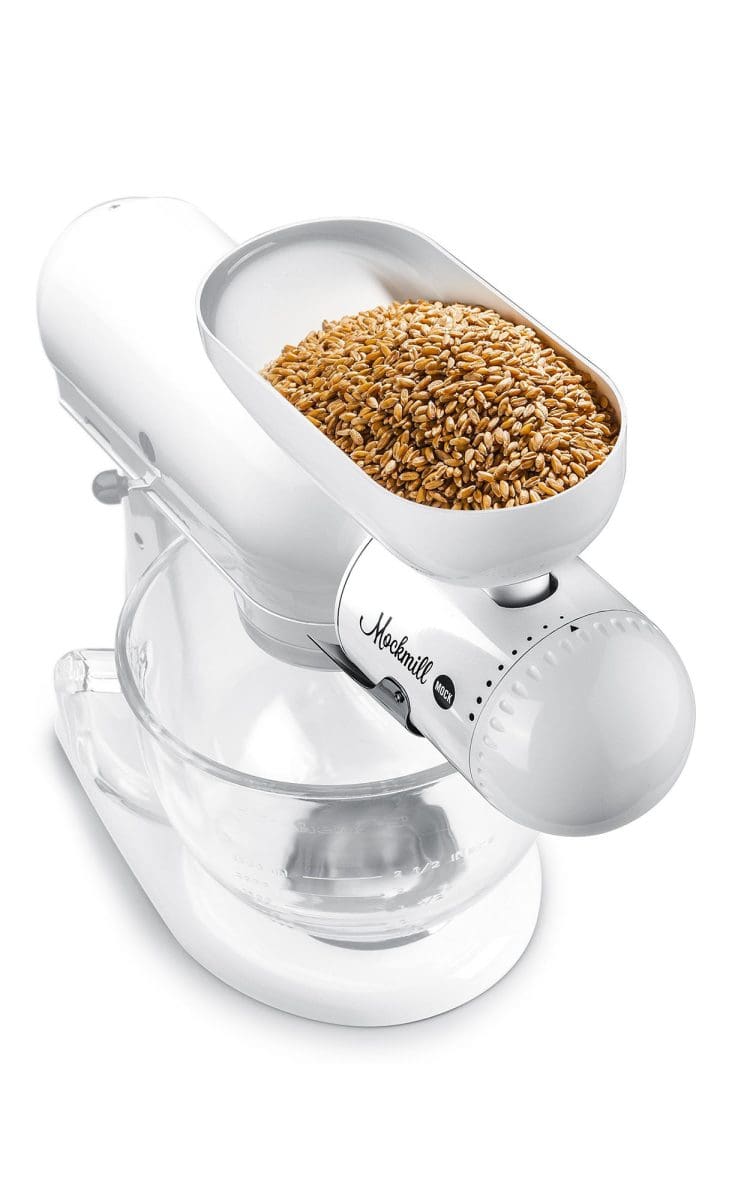 Grain milling attachment grinding wheat into a clear glass bowl, mounted on a kitchen mixer.