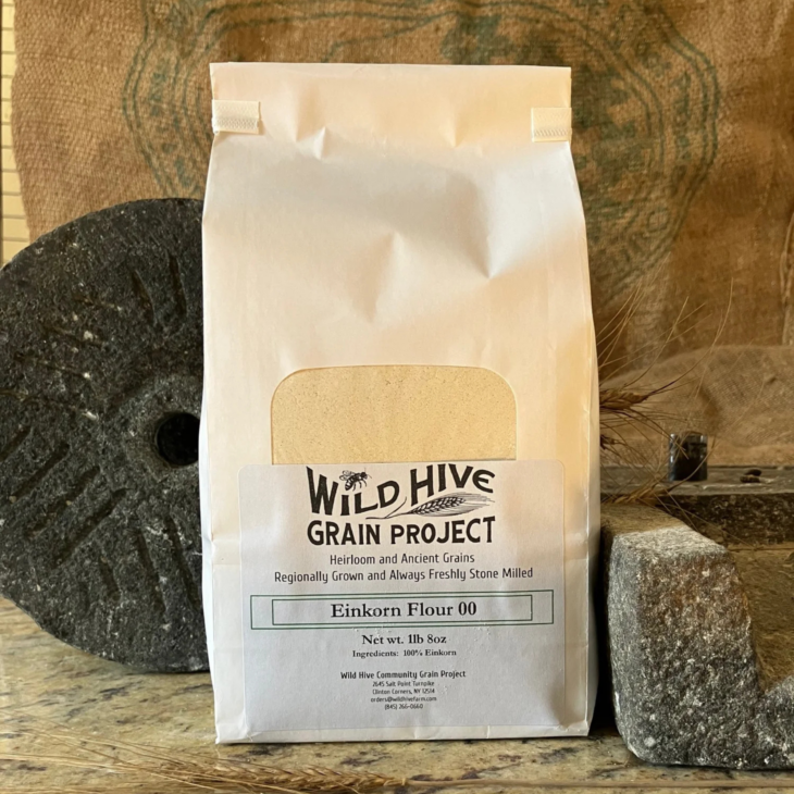 White paper bag of Wild Hive Grain Project Einkorn Flour 00, standing beside a stone mill wheel.